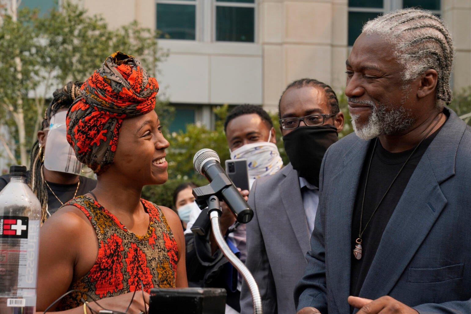 Outdoor press event: a woman in a patterned headwrap smiles beside a microphone while speaking with a man in a blazer with braided hair; masked onlookers gather behind in front of an office building.