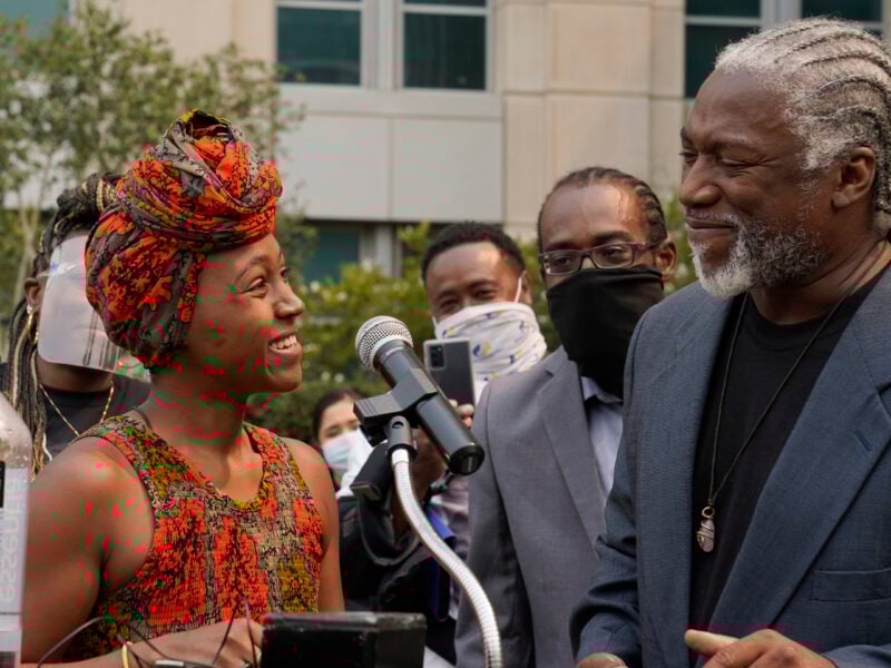Outdoor press event: a woman in a patterned headwrap smiles beside a microphone while speaking with a man in a blazer with braided hair; masked onlookers gather behind in front of an office building.