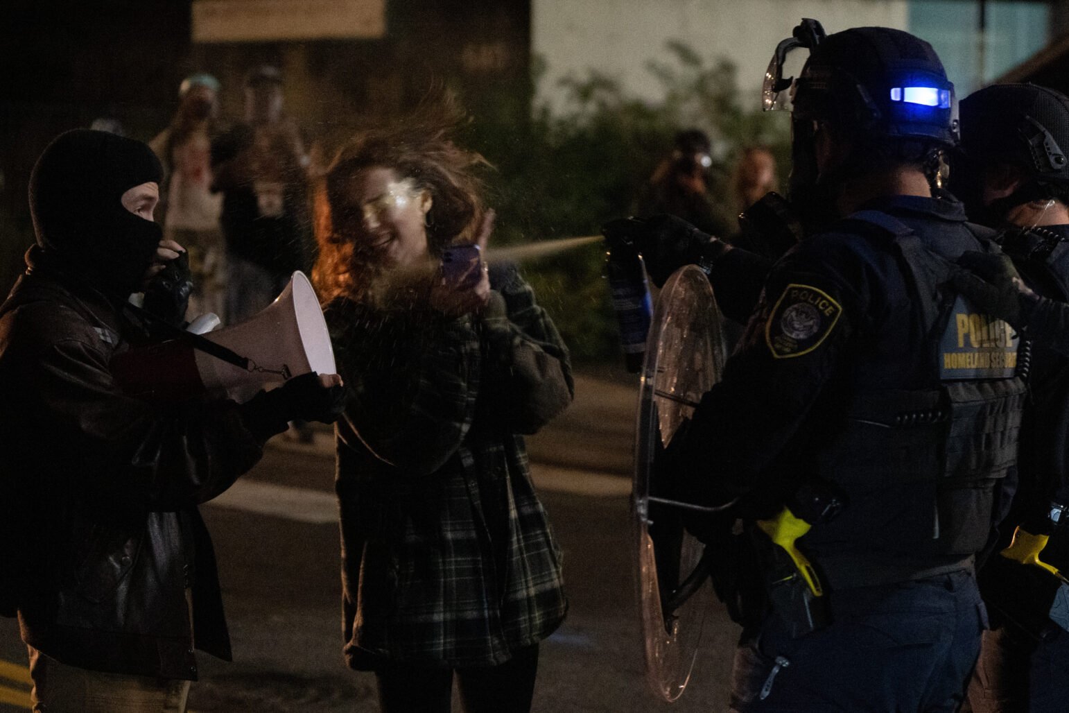 Nighttime street scene where two civilians face police in riot gear—one masked person holds a megaphone while another, wearing a plaid jacket and holding a phone, is struck by a visible mist of spray from an officer’s canister beside a clear riot shield.