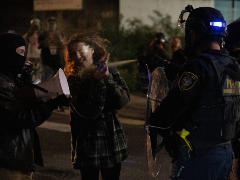 Nighttime street scene where two civilians face police in riot gear—one masked person holds a megaphone while another, wearing a plaid jacket and holding a phone, is struck by a visible mist of spray from an officer’s canister beside a clear riot shield.