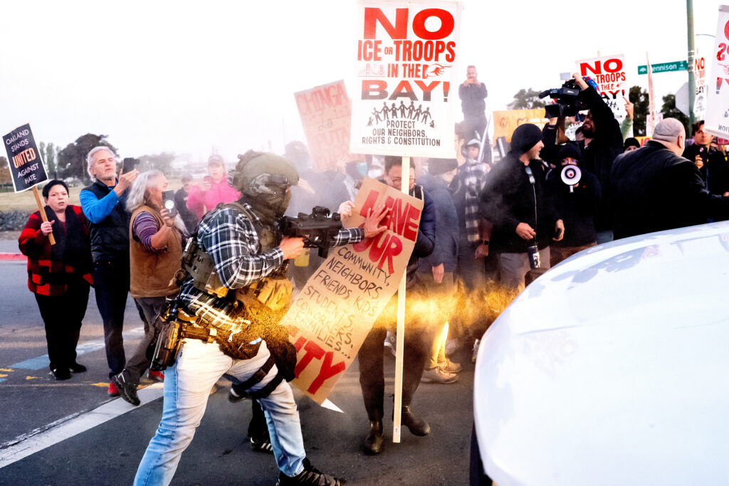 Street protest where a person in tactical gear and a plaid shirt points a rifle and shoves against a protester’s sign amid a burst of spray or smoke; surrounding demonstrators hold placards including NO ICE OR TROOPS IN THE BAY! and Leave our community… while media with cameras and a megaphone look on; a white car fills the right foreground.