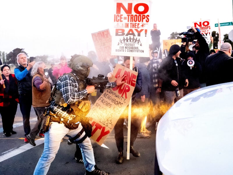 Street protest where a person in tactical gear and a plaid shirt points a rifle and shoves against a protester’s sign amid a burst of spray or smoke; surrounding demonstrators hold placards including NO ICE OR TROOPS IN THE BAY! and Leave our community… while media with cameras and a megaphone look on; a white car fills the right foreground.