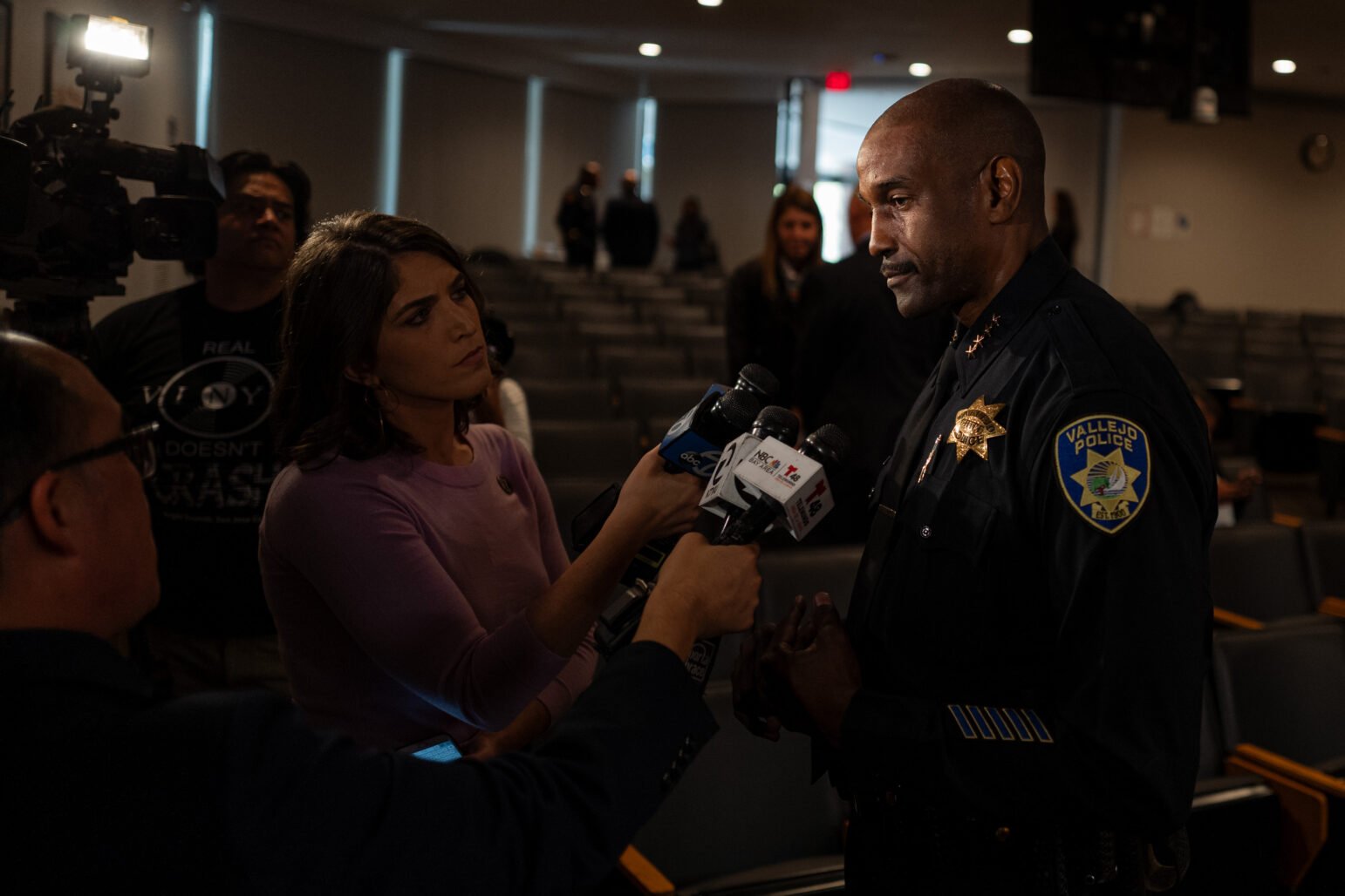 Police chief in a Vallejo Police Department uniform speaks to reporters holding microphones in a city council chambers, with TV cameras and other people in the background.