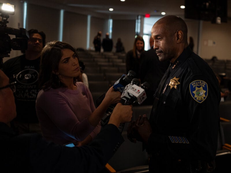 Police chief in a Vallejo Police Department uniform speaks to reporters holding microphones in a city council chambers, with TV cameras and other people in the background.
