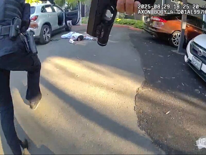 Body-camera view of a police encounter in a parking lot: an officer’s handgun in the foreground, another officer at left, and a person in white clothing lying face-down near an open SUV. Evening shadows and a timestamp overlay the scene.