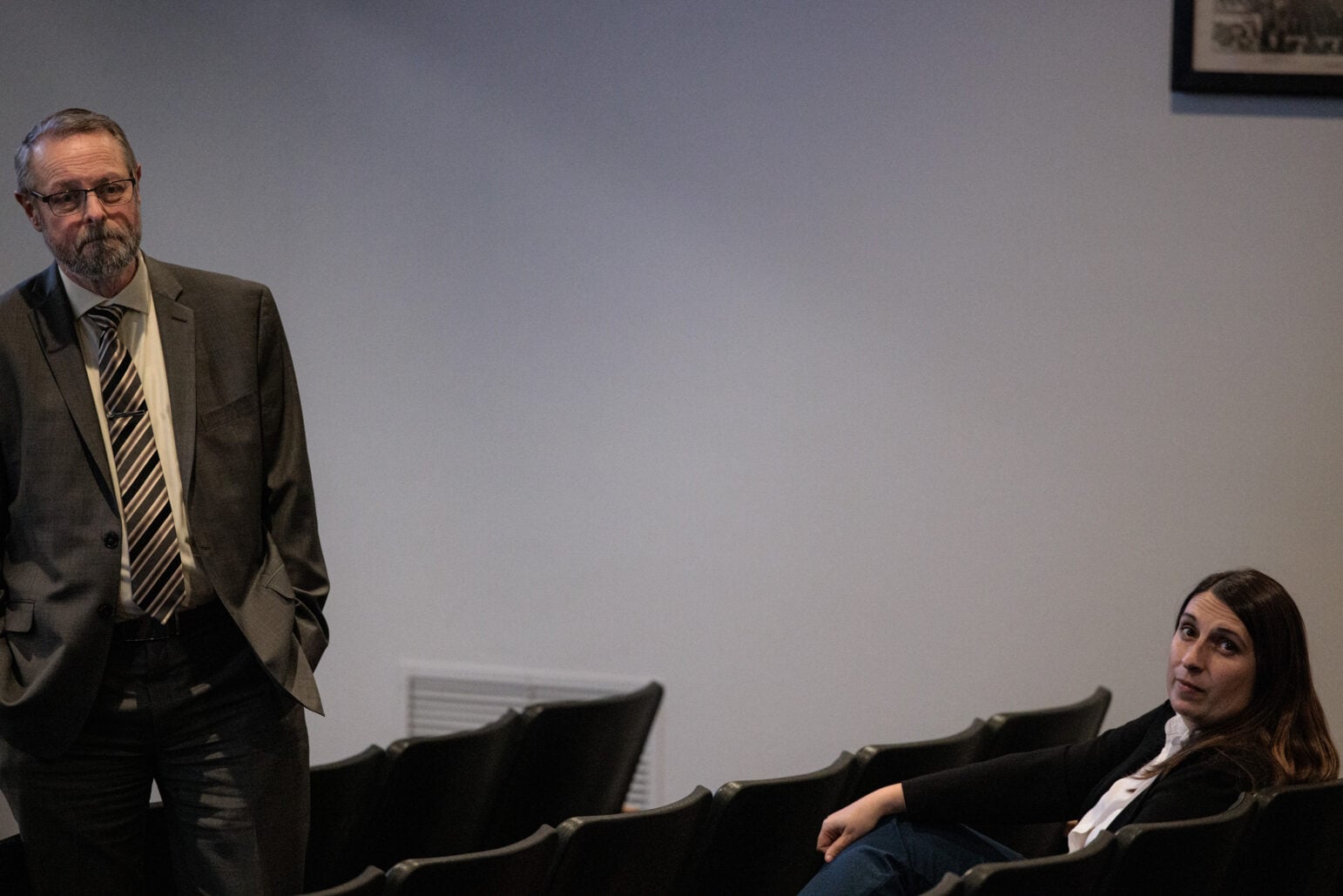 A man in a gray suit and striped tie stands with his hands clasped while a woman with long brown hair in a black blazer sits nearby in a row of empty seats during a city council meeting.