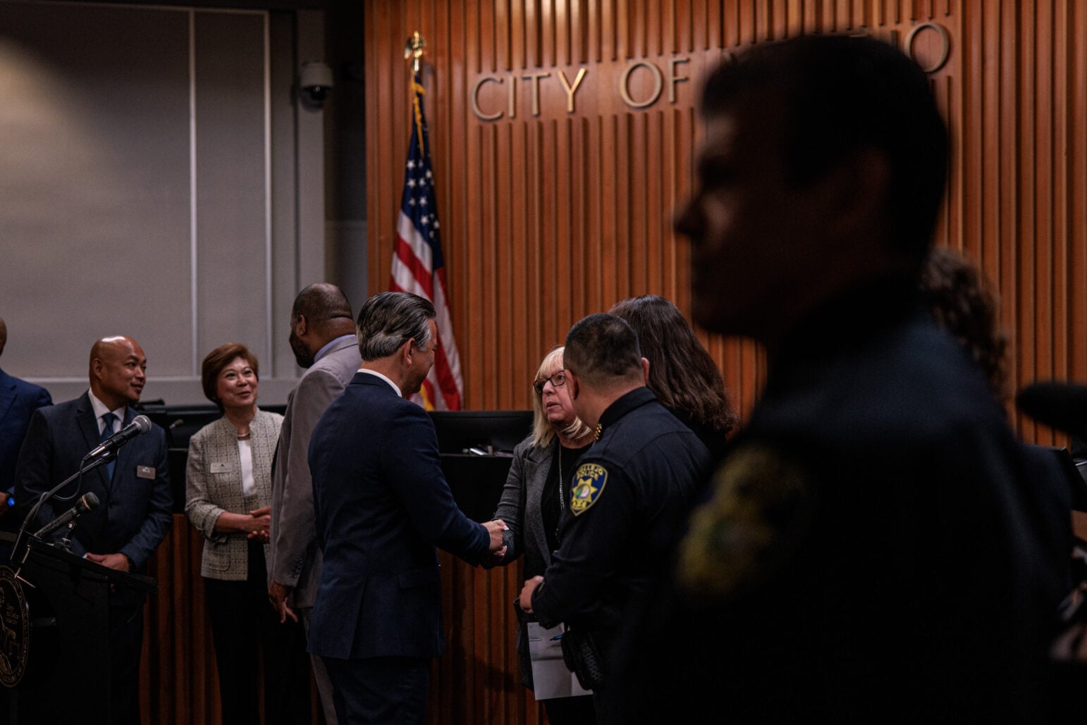 California Attorney General Rob Bonta, in a dark blue suit, shakes hands with Vallejo City Attorney Veronica Nebb inside city council chambers, with other officials and an American flag visible in the background.