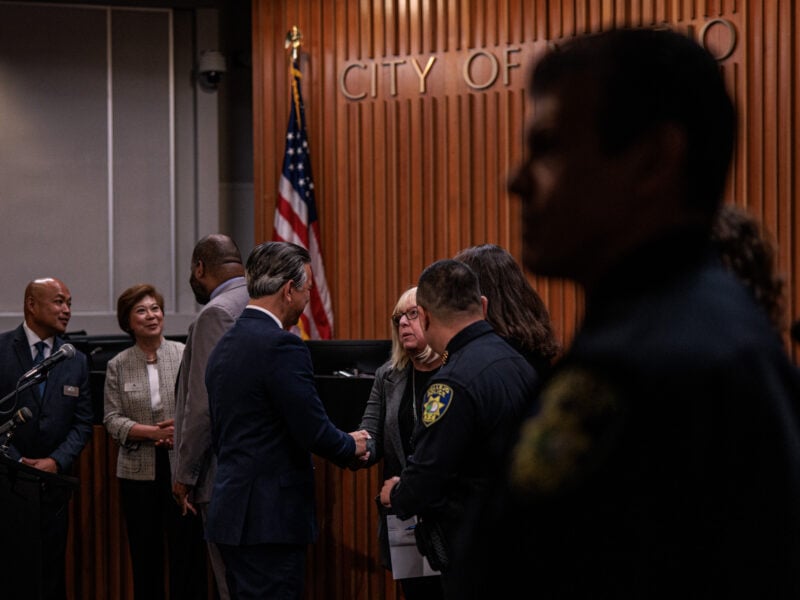 California Attorney General Rob Bonta, in a dark blue suit, shakes hands with Vallejo City Attorney Veronica Nebb inside city council chambers, with other officials and an American flag visible in the background.
