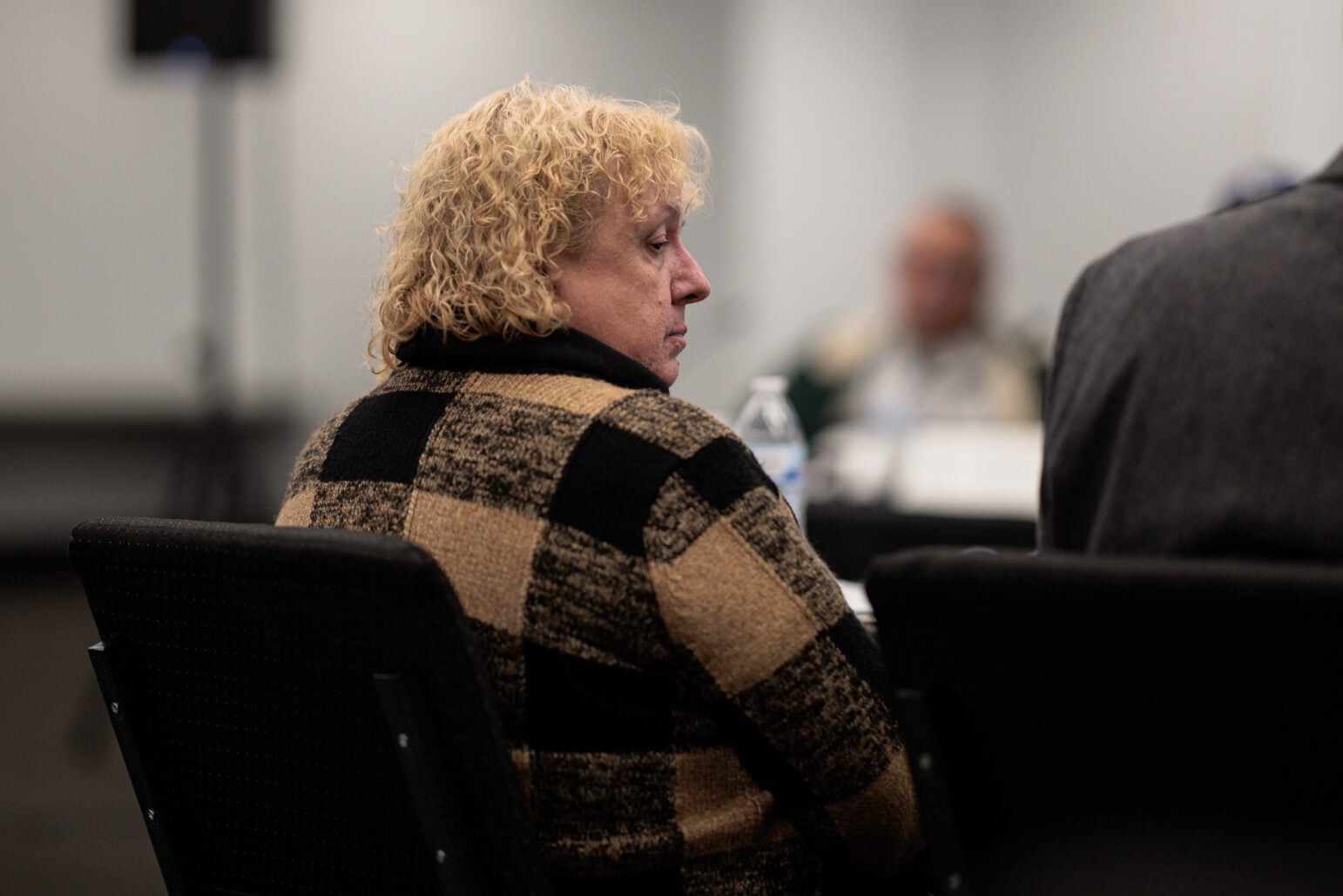 Profile view of a woman with curly blonde hair wearing a black and tan checkered sweater, seated in a meeting room.
